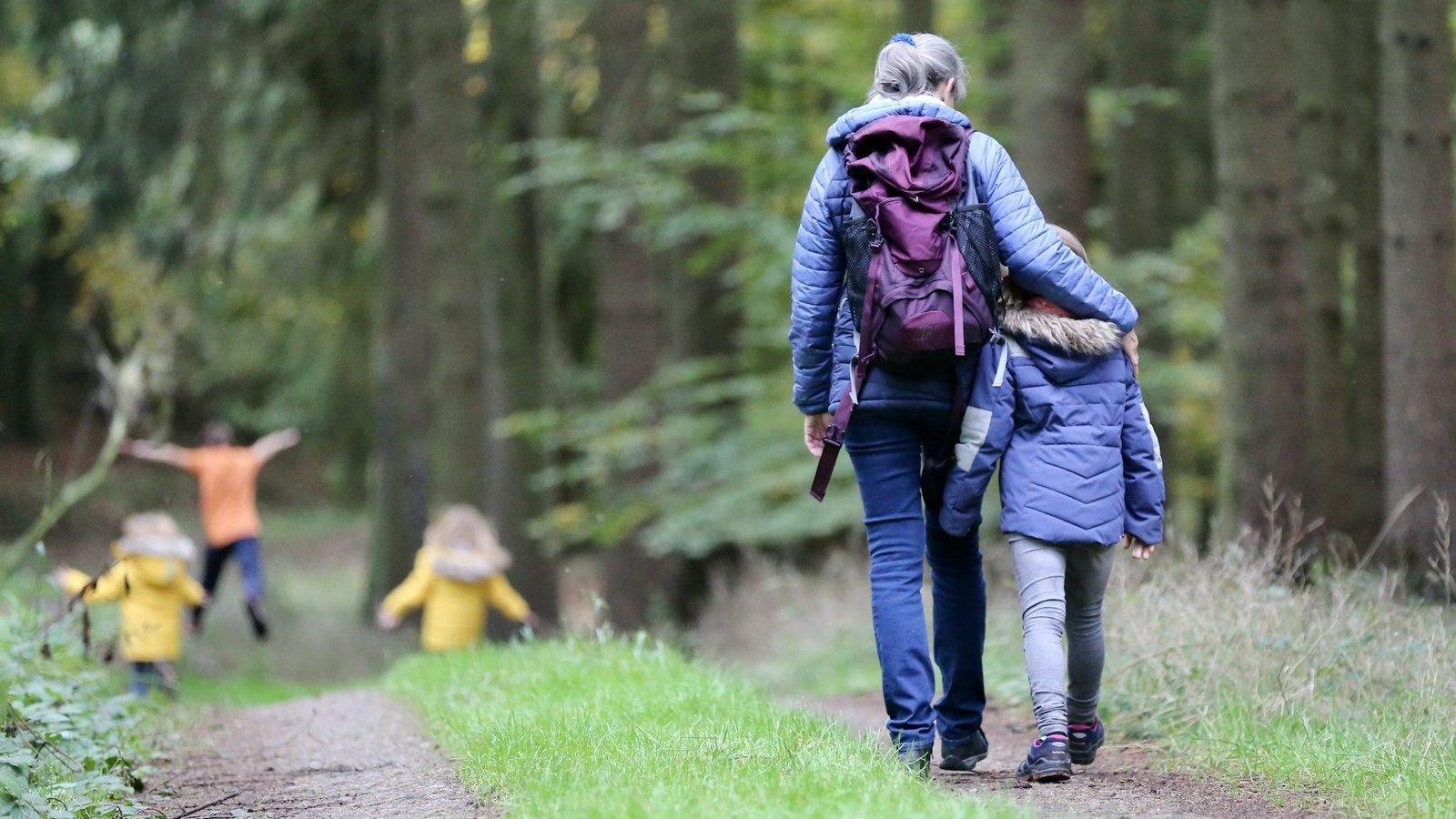 Eine Frau hat die Schulter um ein Kind gelegt. Sie spazieren auf einem Weg durch einen Wald. Unscharf im Vordergrund sind weitere Kinder vorne zu sehen.