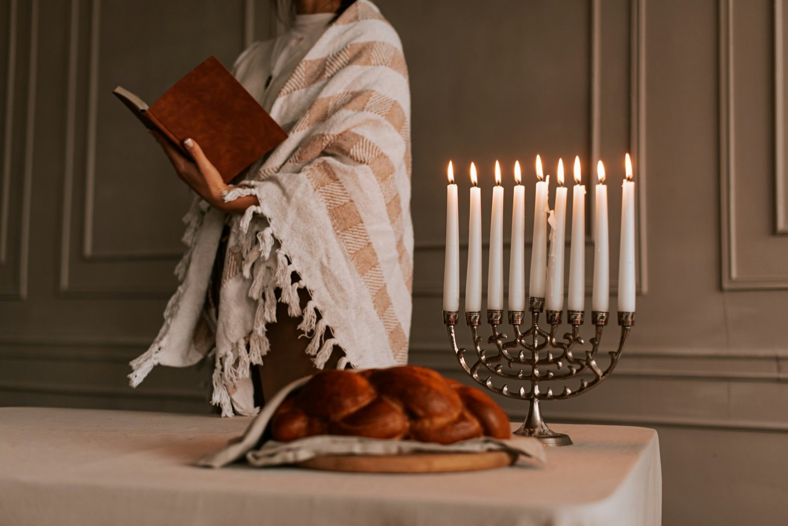 Eine Person steht mit einem gestreiften Gebetstuch um die Schultern und einem Buch in der Hand hinter einem Tisch auf dem eine Menora (siebenarmiger Leuchter) und Challa (traditionelles jüdisches Weißbrot) stehen.