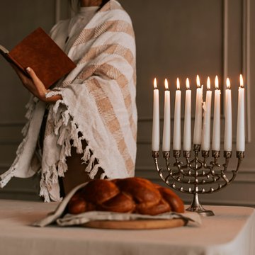 Eine Person steht mit einem gestreiften Gebetstuch um die Schultern und einem Buch in der Hand hinter einem Tisch auf dem eine Menora (siebenarmiger Leuchter) und Challa (traditionelles jüdisches Weißbrot) stehen.