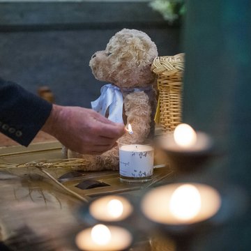 Mensch zündet eine Kerze an, daneben sitzt ein Teddybär auf dem Boden und weitere Kerzen.