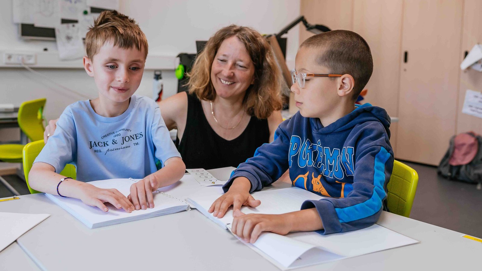 Zwei blinde Schüler sitzen mit ihrer Lehrerin im Klassenzimmer und lesen die Brailleschrift.