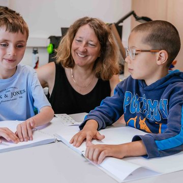 Zwei blinde Schüler sitzen mit ihrer Lehrerin im Klassenzimmer und lesen die Brailleschrift.
