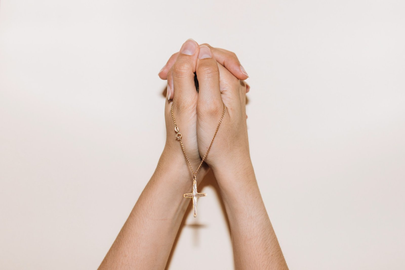 Gefaltete Hände einer weißen Person halten eine goldene Kette mit einem Kreuz-Anhänger.