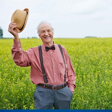 Ein älterer Mann steht auf einem Feld und hebt seinen Hut zum Gruß.