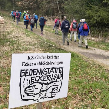 Im Vordergrund eine Tafel auf der Gedenkstätte steht, rechts daneben ein Weg auf dem Wanderer laufen.