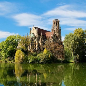 Die Johannes Kirche in Stuttgart ist auf dem Foto zu sehen. Sie liegt am Feuersee und ist umgeben von grünen Bäumen.