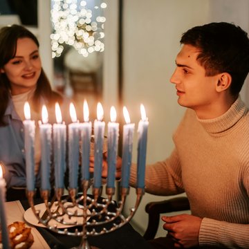 Junger Mann und junge Frau sitzen an einem Tisch, Kerzen an einem siebenarmigen Leuchter brennen.