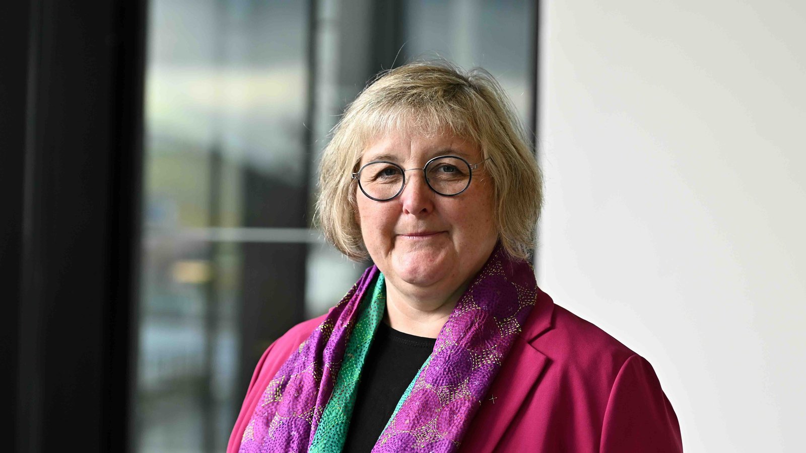 Portraitfoto zeigt die Bischöfin der Landeskirche in Baden, Heike Springhart. Sie trägt ein schwarzes Shirt und einen pinken Blazer sowie einen bunten Schal. Sie hat blonde Haare, trägt eine Brille und lächelt in die Kamera.