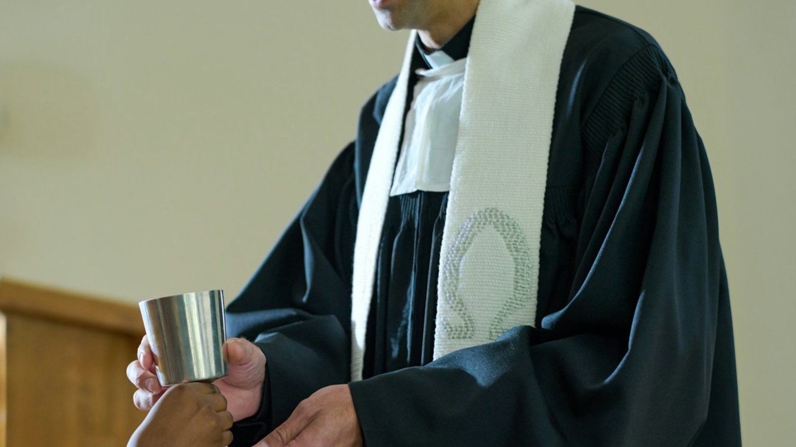 Ein Mann in schwarzem Talar mit weißem Beffchen und weißer Stola reicht einer Frau, die vor ihm steht einen Kelch. 