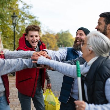 Sechs Menschen (jung und alt) stehen in einem Halbkreis und halten alle einen Arm nach vorne, sodass sich alle Hände berühren. Eine ältere Frau mit grauen Haaren vorne rechts hat einen Stab in der Hand.