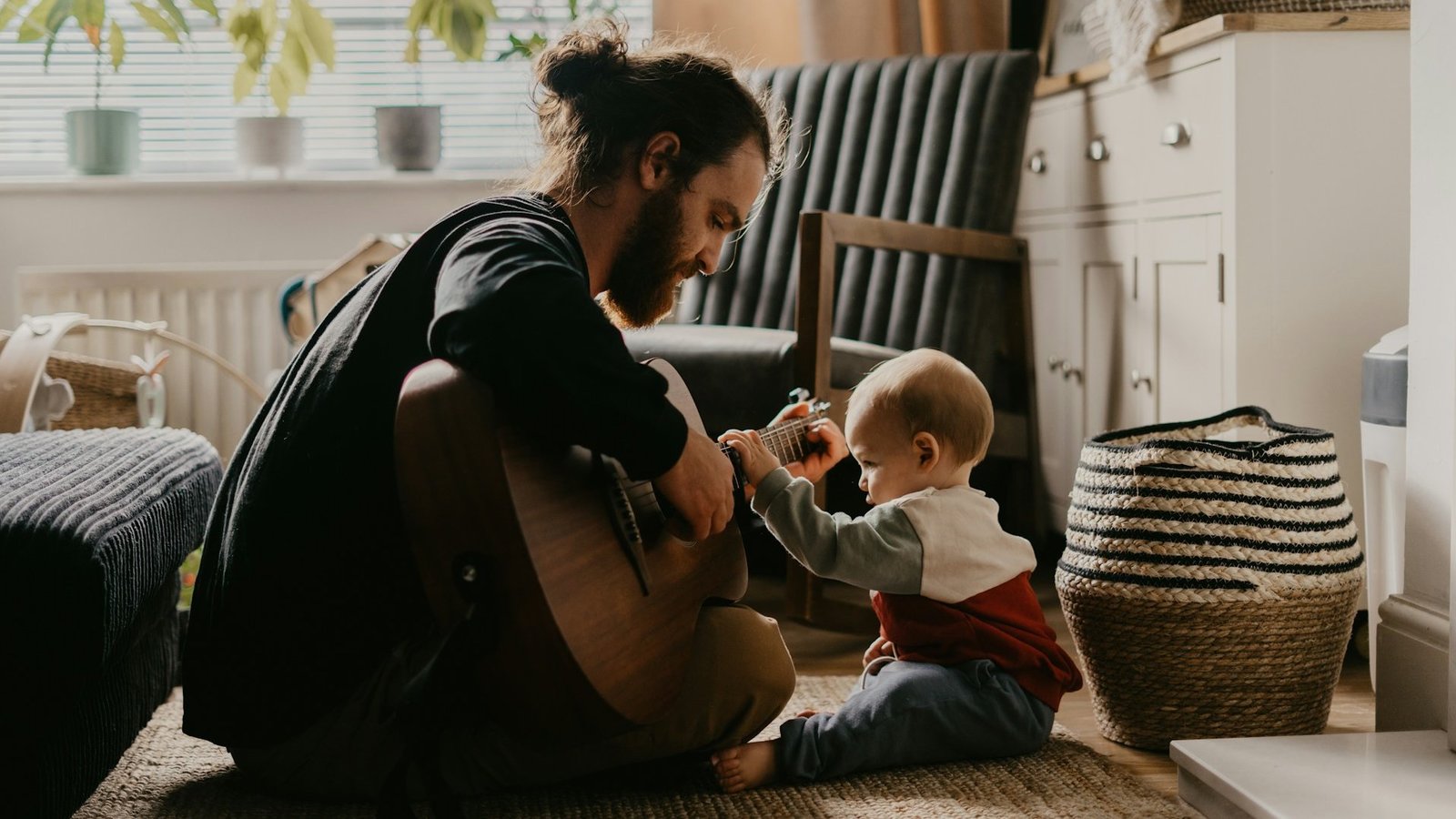 Ein Mann sitzt in einem Zimmer auf dem Boden und spielt eine Gitarre. Ihm gegenüber sitzt ein Baby und hat eine Hand am Hals der Gitarre.