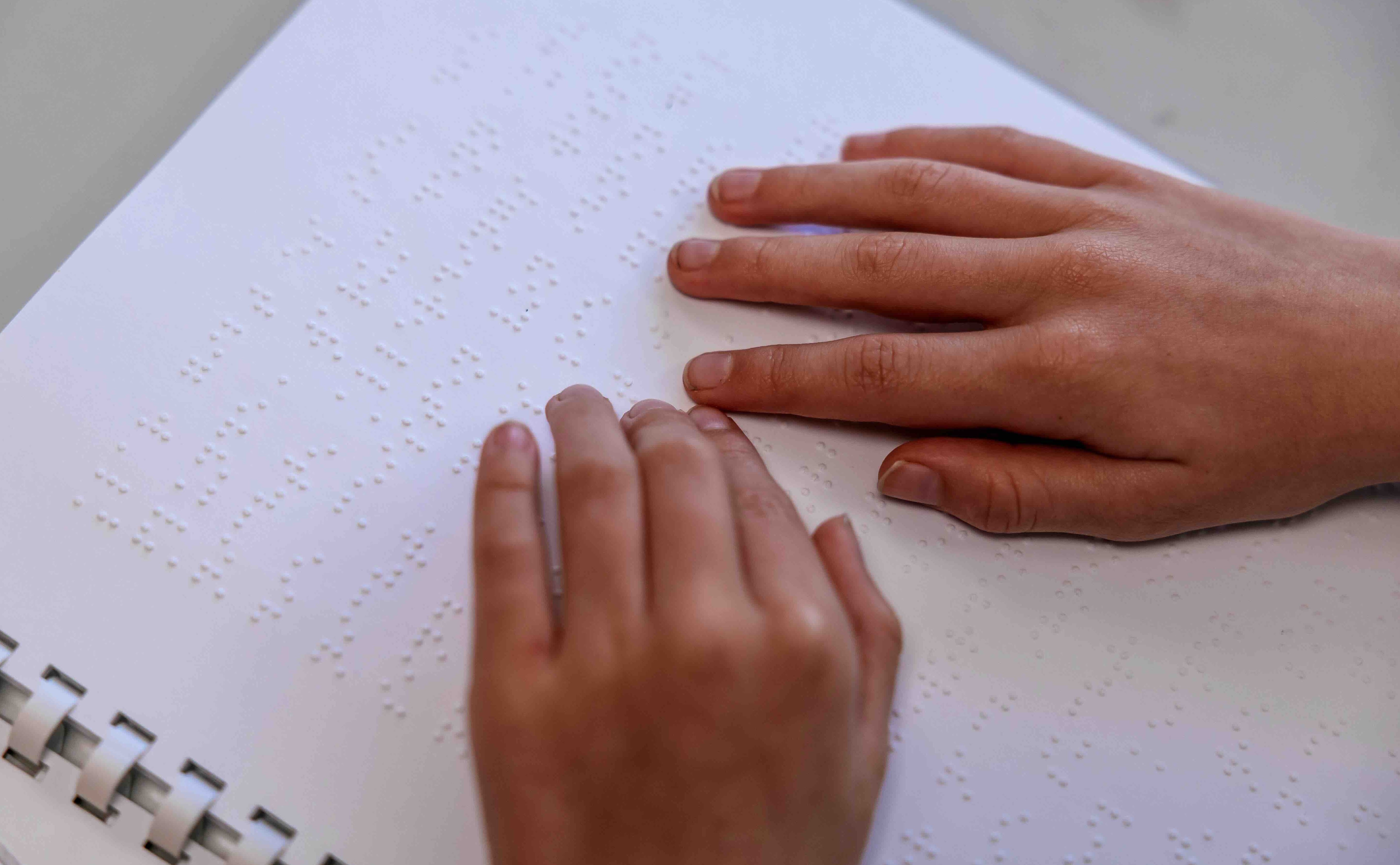 Die Aufnahme zeigt ein weißes Blatt mit der Brailleschrift und zwei Hände, die die Schrift ertasten.