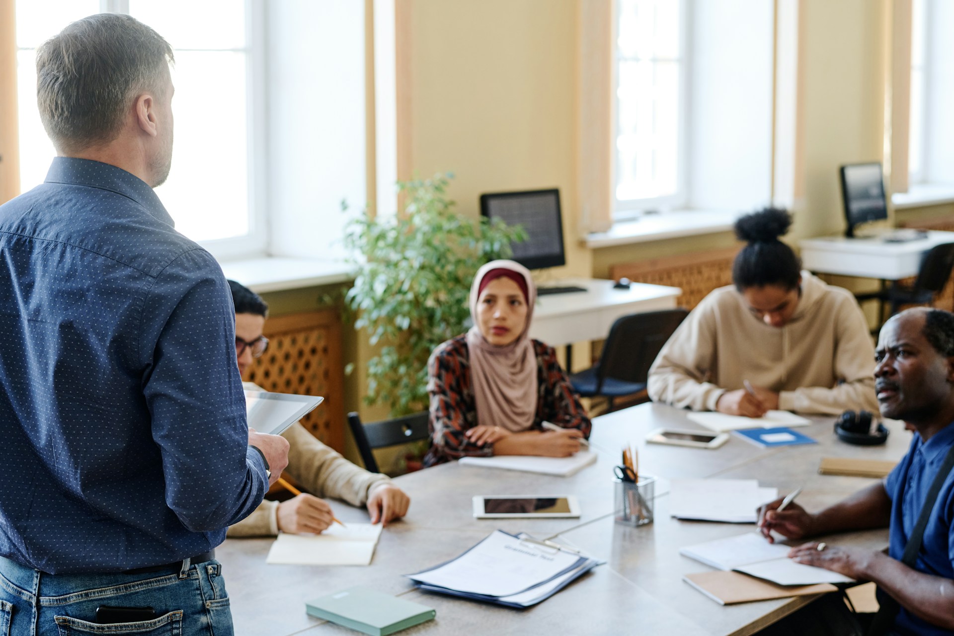 Eine Gruppe von Menschen aus verschiedenen Ländern sitzt gemeinsam an einem runden Tisch und schreibt auf Blätter. Links vorne im Bild steht ein Mann, der den Menschen etwas beizubringen scheint.
