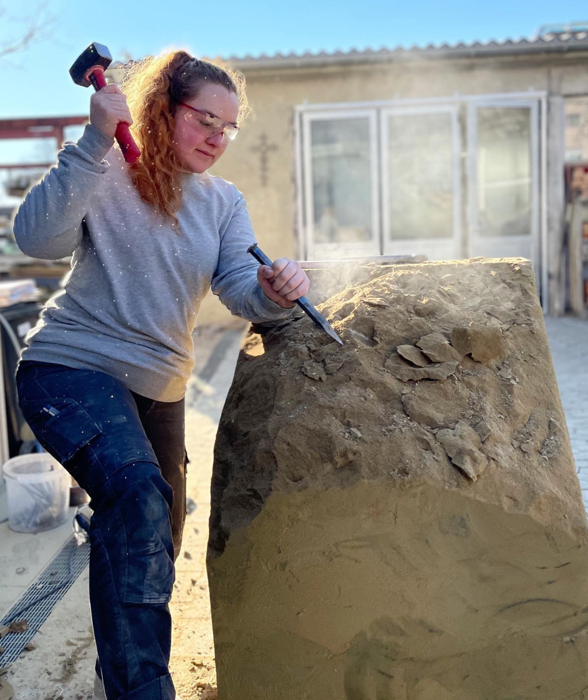 Das Bild zeigt Luisa Lüttich bei der Bearbeitung eines hüfthohen Steines mit Hammer und Meisel.