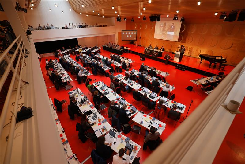 Synode tagt im Hospitalhof. An Tischen sitzen viele Menschen, auf einer Bühne vorne steht ein Redner, der Fußboden ist rot. 