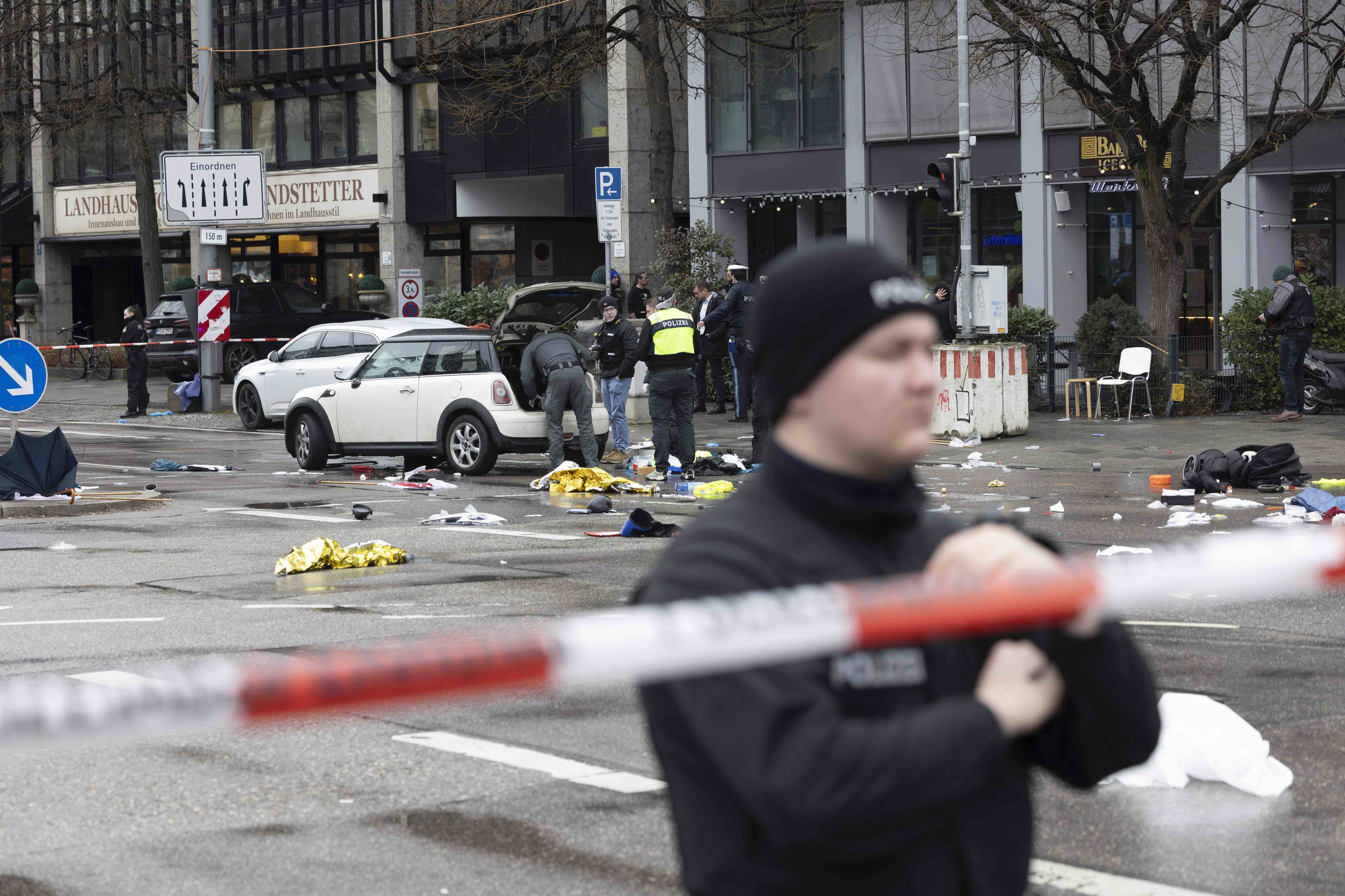 Das Bild zeigt den Tatort nach dem Anschlag in München am 13. Februar 2025. Polizeiabsperrband und ein Polizist im Vordergrund. Im Hintergrund ist das Tatfahrzeug zu sehen, in dem Polizisten in den Kofferraum schauen. 