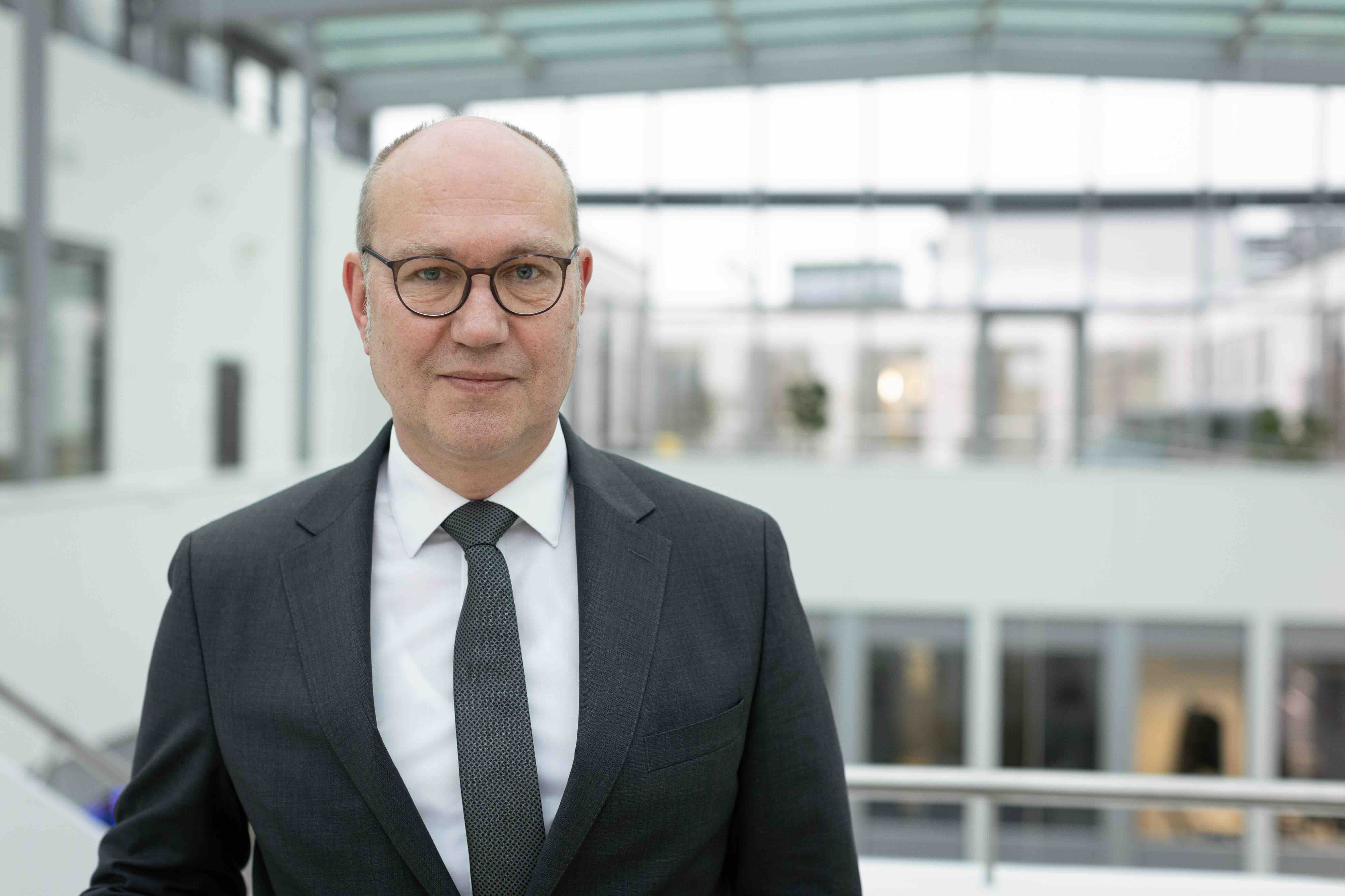 Portraitfoto von Rüdiger Schuch, der Mann trägt einen Anzug und Krawatte und ein weißes Hemd. Außerdem trägt er eine Brille mit dunklem Rand.