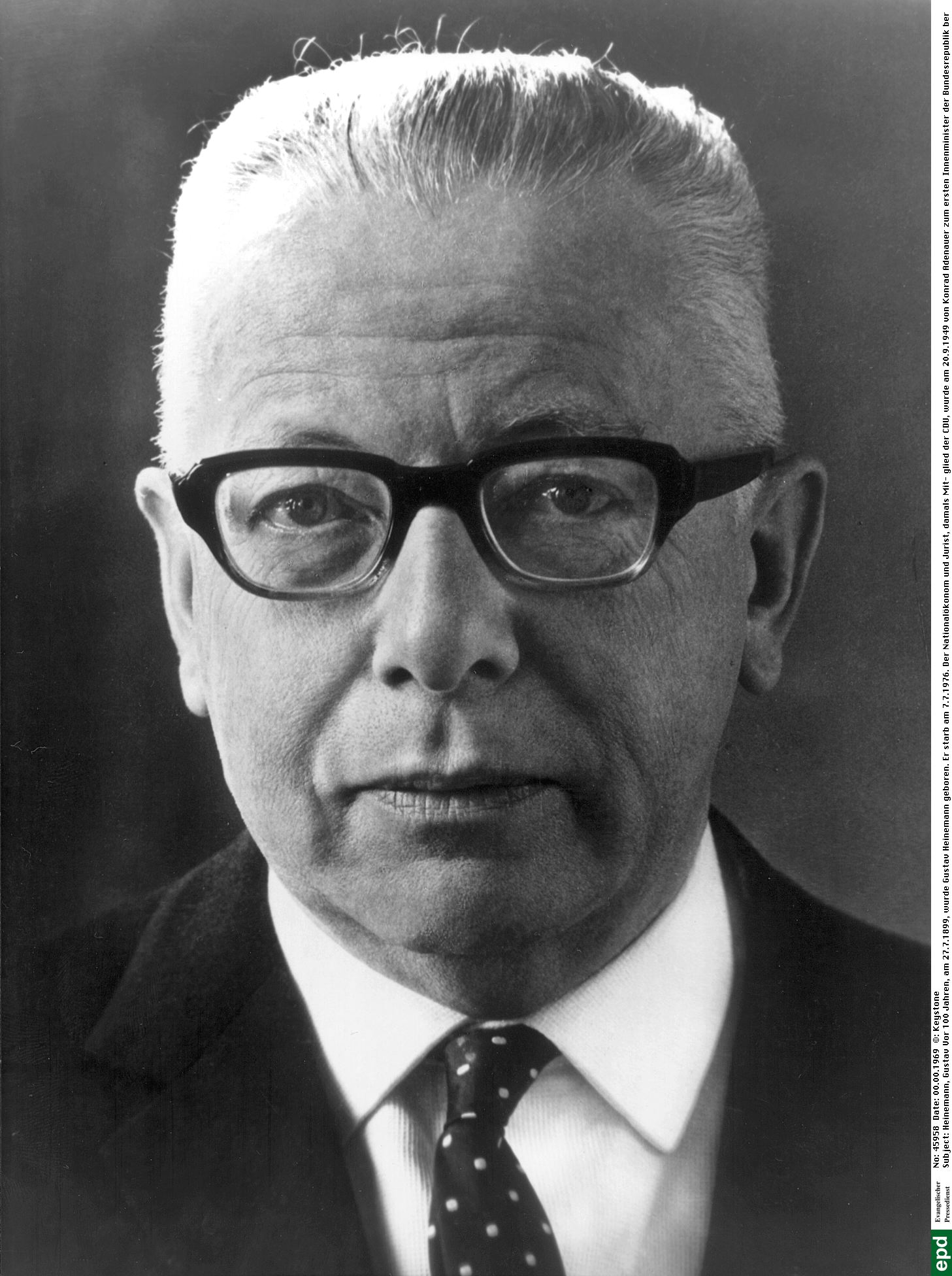 Die schwarz-weiß Fotografie zeigt ein Portrait von Gustav Heinemann. Er trägt ein weißes Hemd, Jackett, Krawatte, eine Kurzhaarfrisur und eine Brille mit dicken dunklen Rändern.