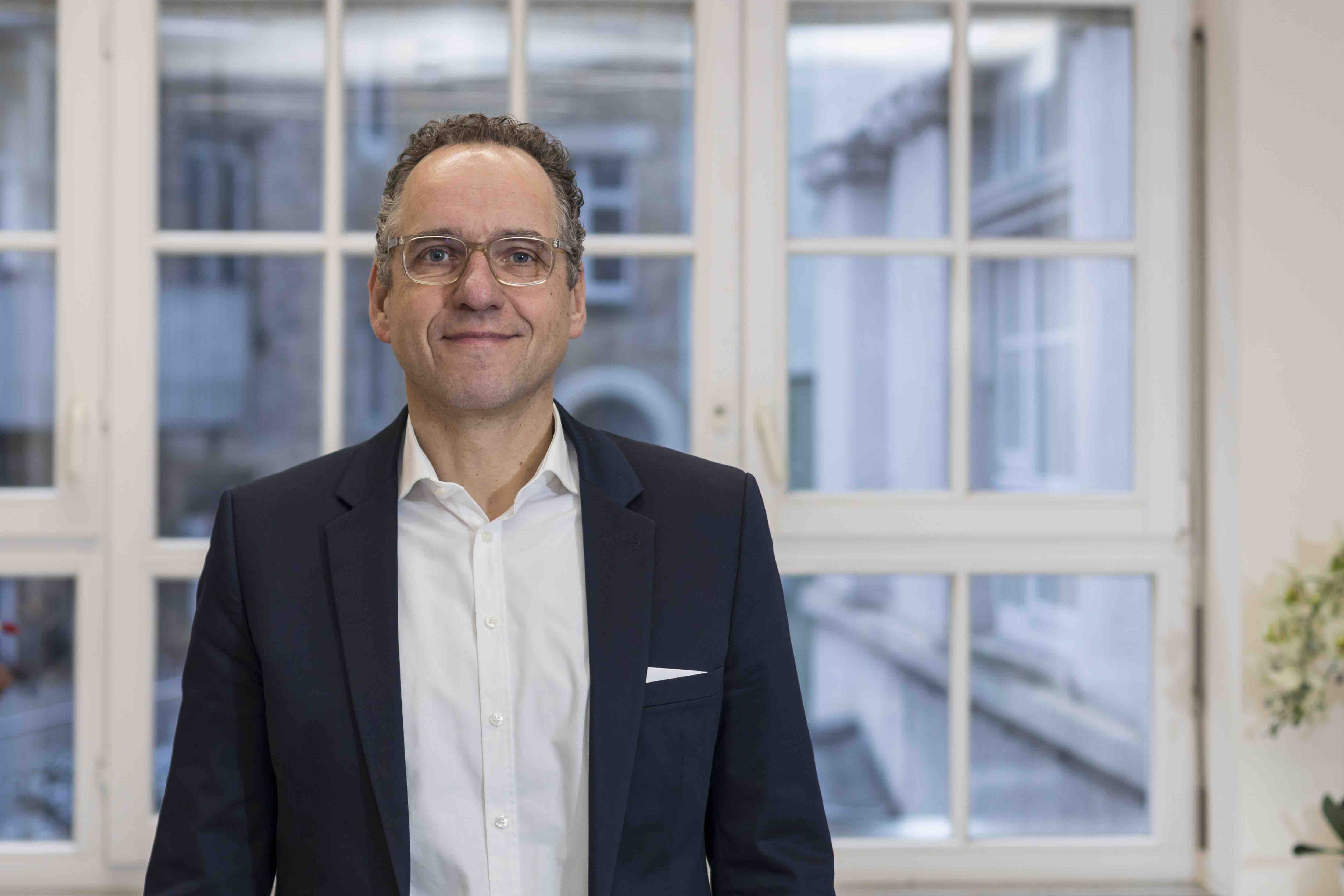 Portrait von Frank Zeithammer. Er trägt ein weißes Hemd, ein graues Jackett und eine Brille. Er hat graue Haare.