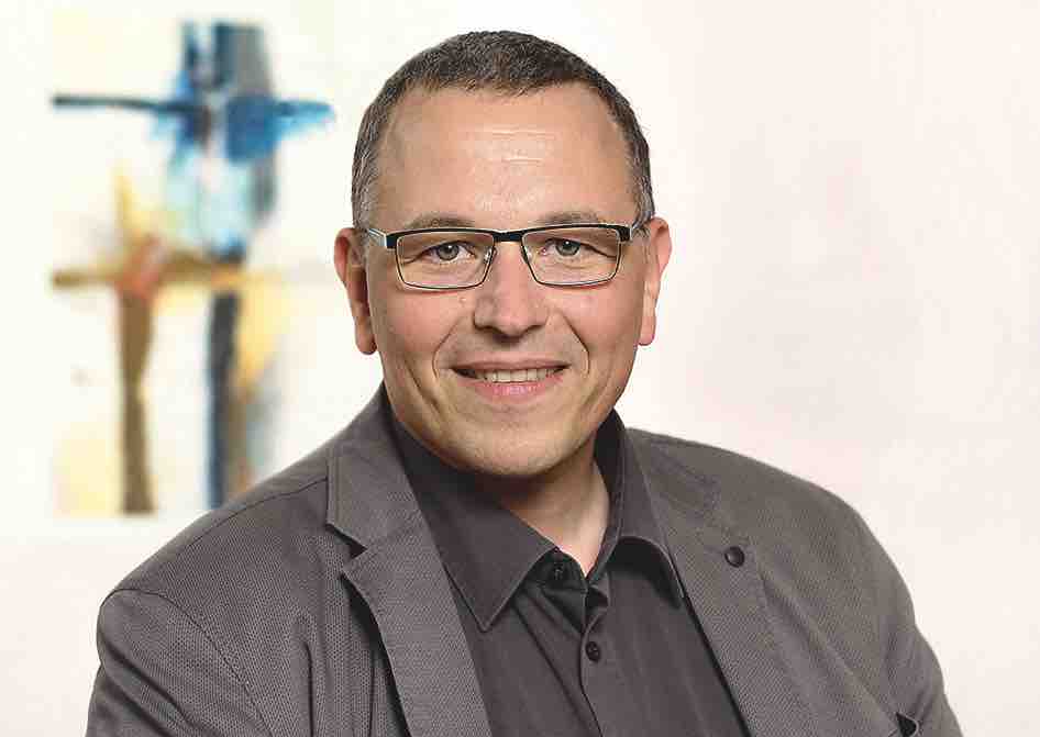 Portrait von Tobias Glawion. Er trägt eine Brille, kurze graue Haare, ein braunes Hemd und ein graues Jackett.