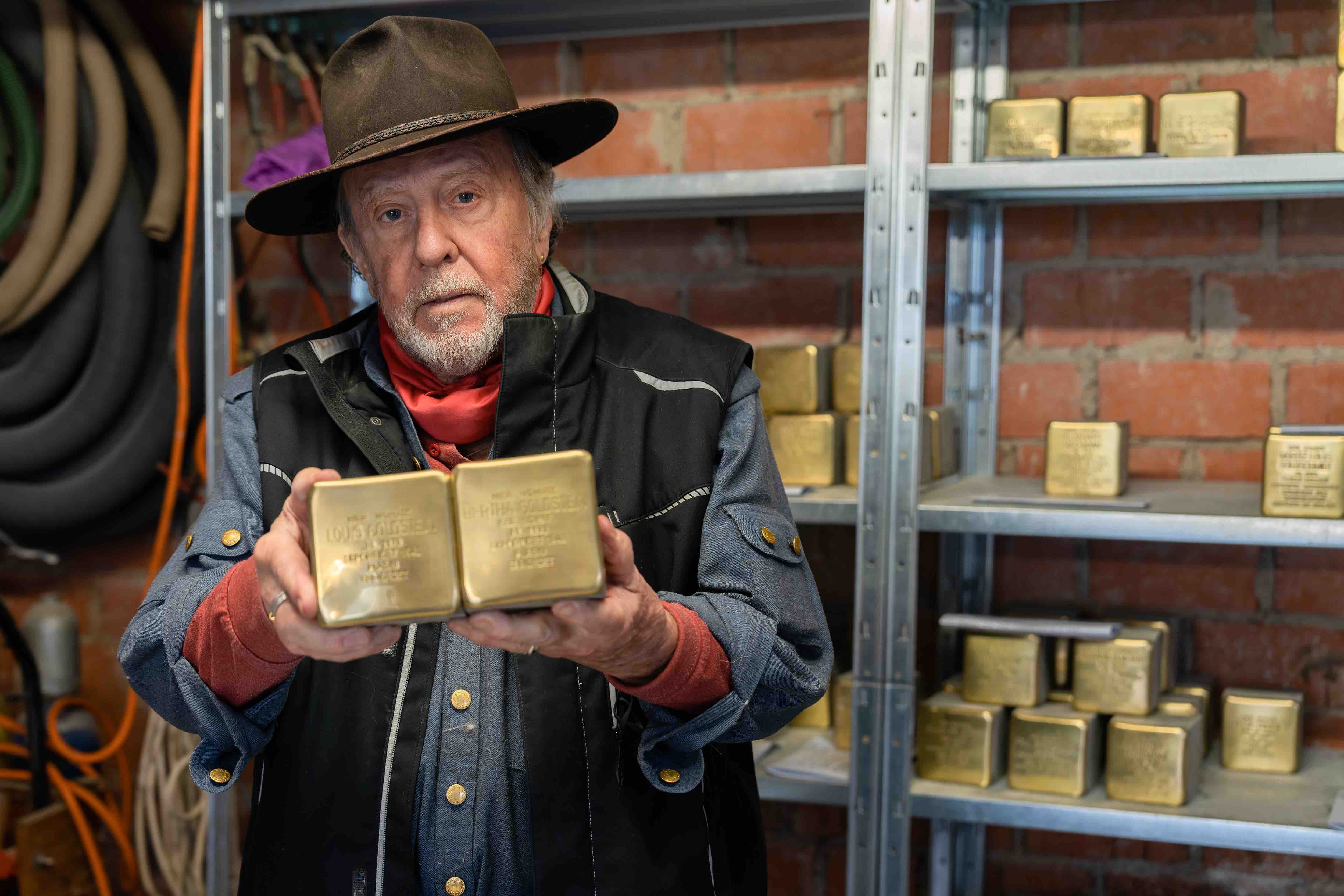 Der Künstler Gunter Demnig hält zwei quadratische goldene "Stolpersteine", die an die Ermordung von Jüdinnen und Juden durch die Nationalsozialisten erinnern, in der Hand. Hinter ihm steht ein Regal in dem weitere "Stolpersteine" stehen