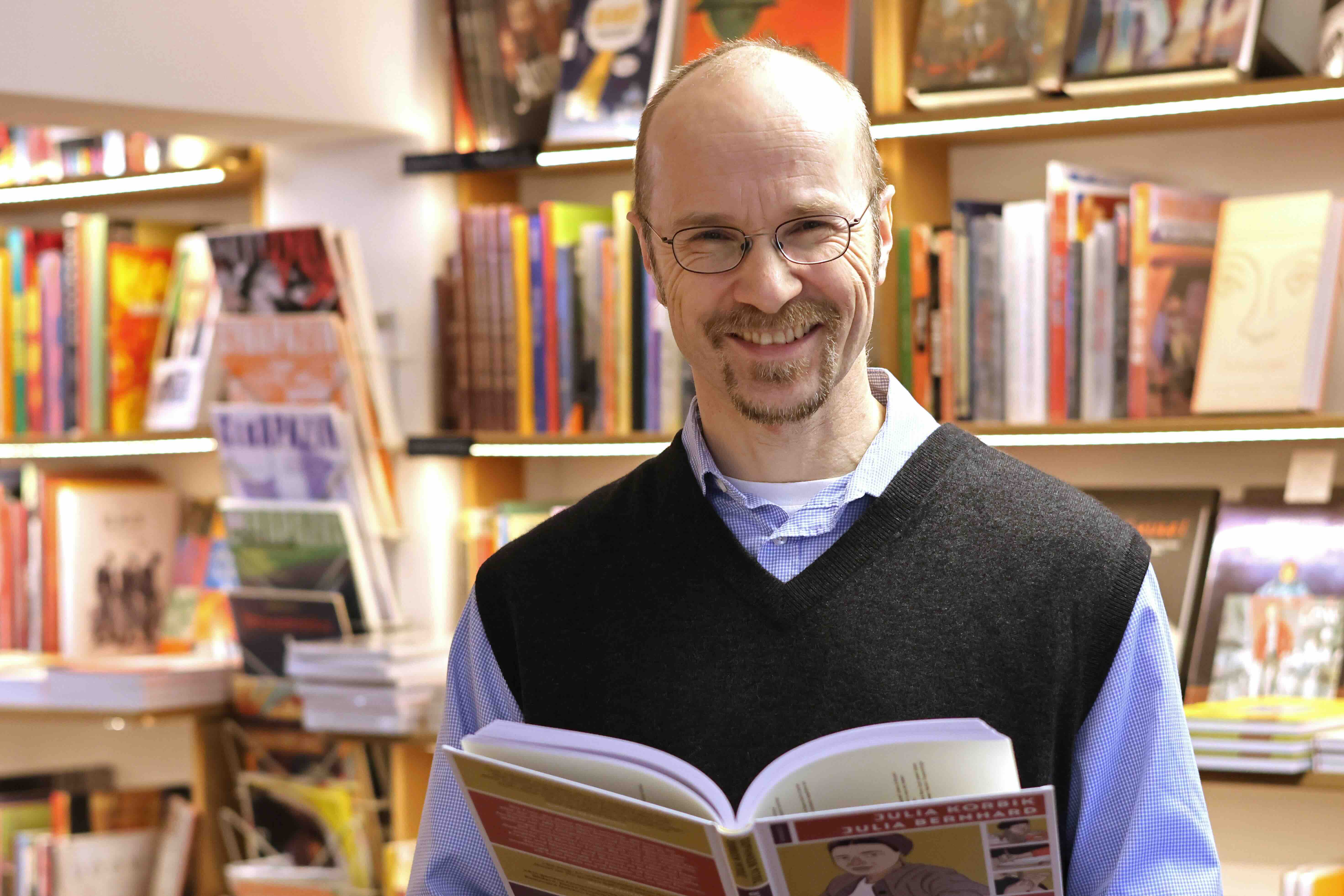 Burkhard Conrad hält ein aufgeschlagenes Buch in den Händen und lächelt in die Kamera. Im Hintergrund sind Bücherregale zu sehen.