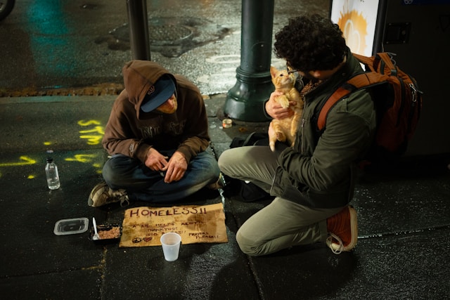 Mann hat eine Katze auf dem Arm und kniet neben einem Obdachlosen