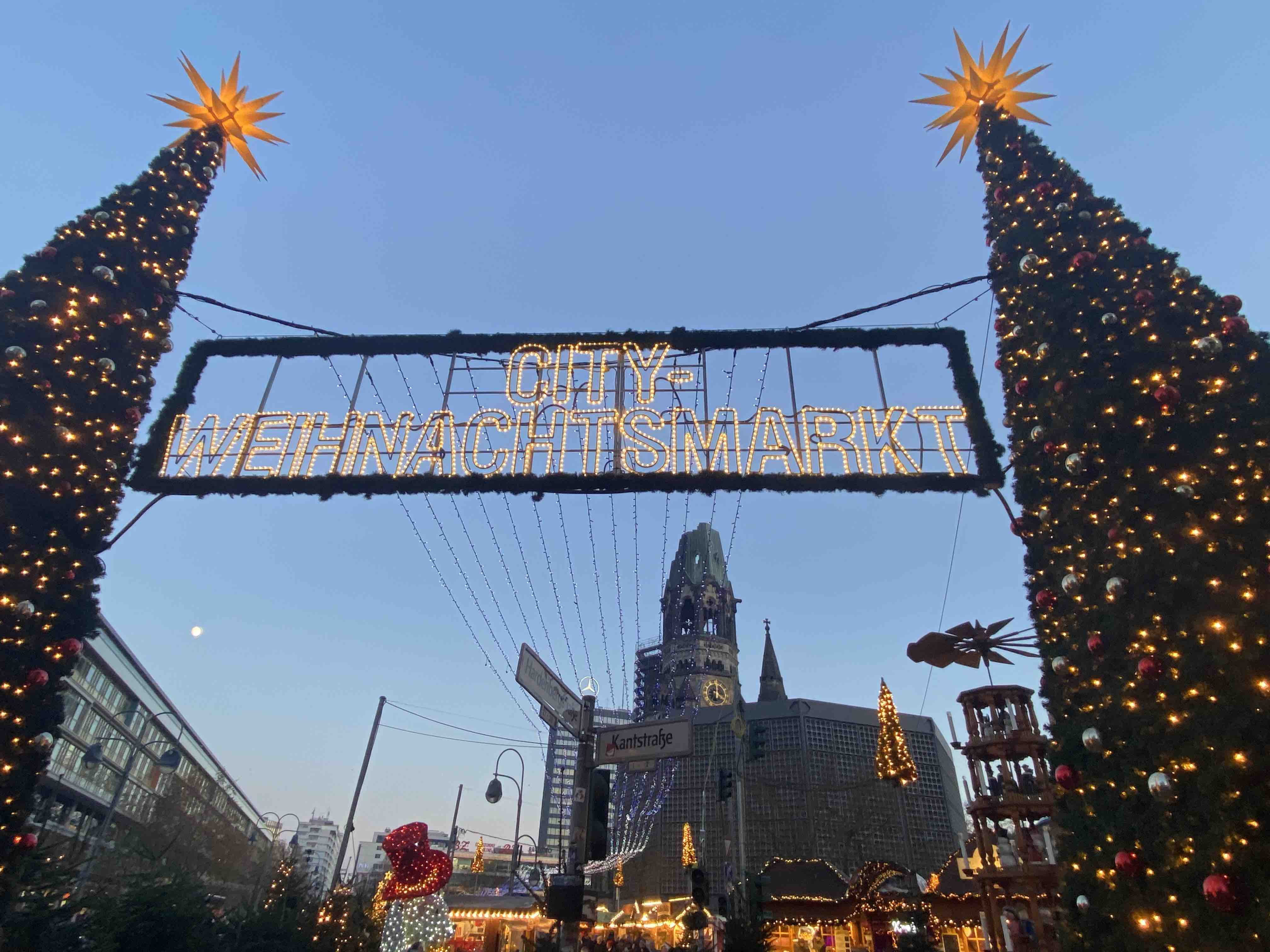 Zwischen zwei mit Lichtern und Kugeln geschmückten Christbäumen ist ein Banner mit Leuchtschrift gespannt auf dem steht: "CITY-WEIHNACHTSMARKT". Im Hintergrund sind Gebäude zu sehen. Das Bild zeigt den Weihnachtsmarkt am Berliner Breitscheidplatz.