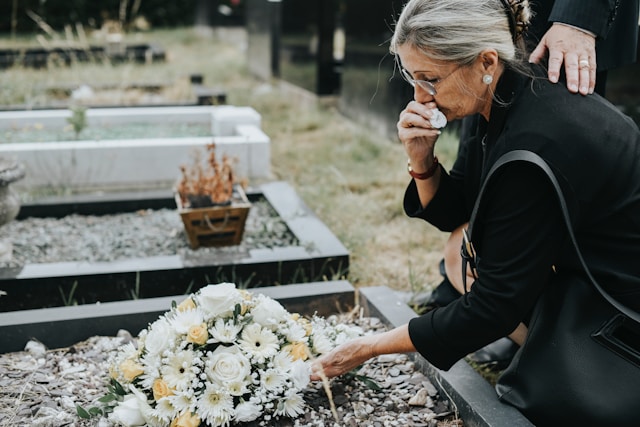 Frau kniet vor Grab auf dem Blumen liegen und hält sich ein Taschentuch unter die Nase.