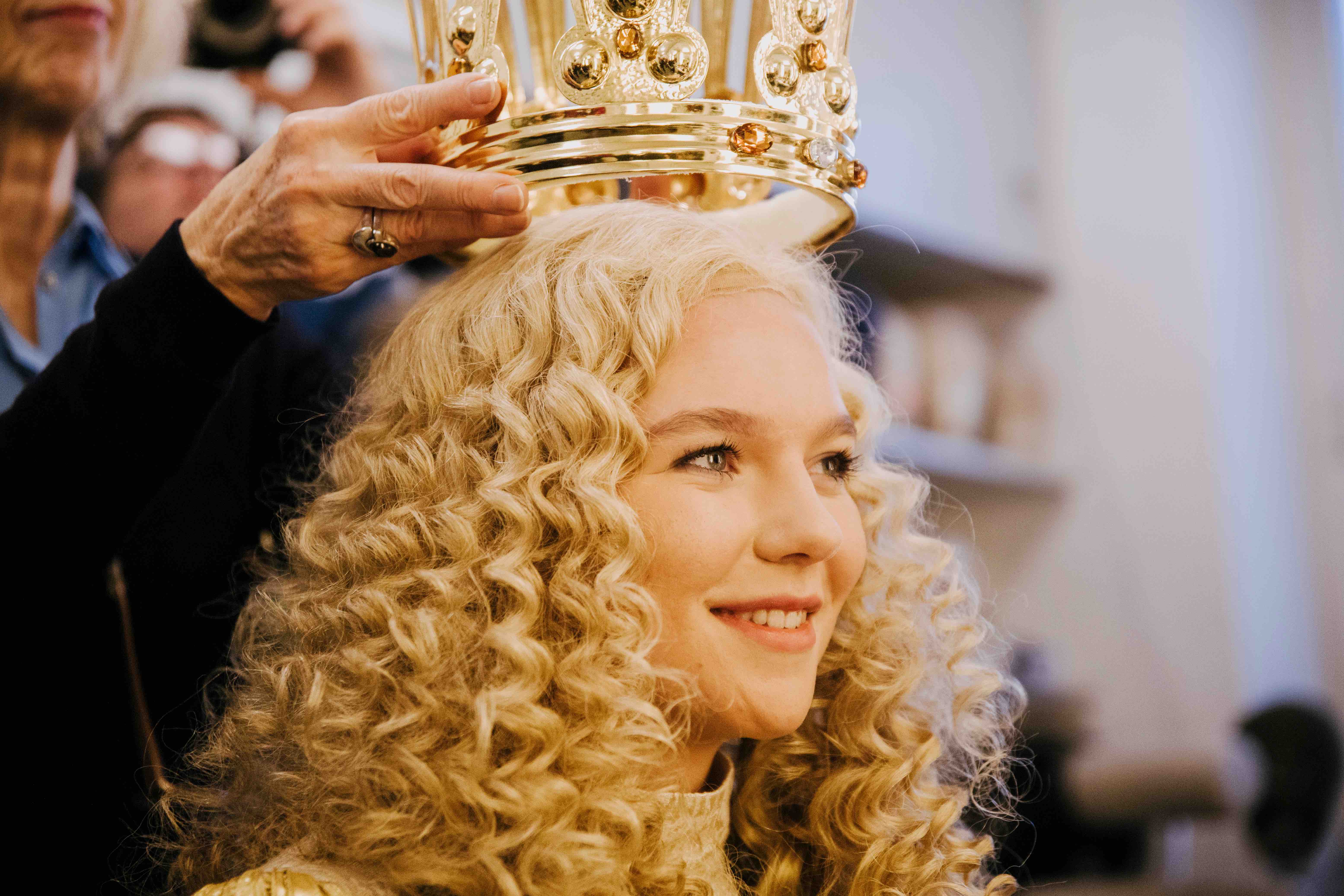 Die Krönung des Nürnberger Christkindes. Die junge Frau hat blonde lockige Haare. Eine Person setzt ihr eine goldene Krone auf.
