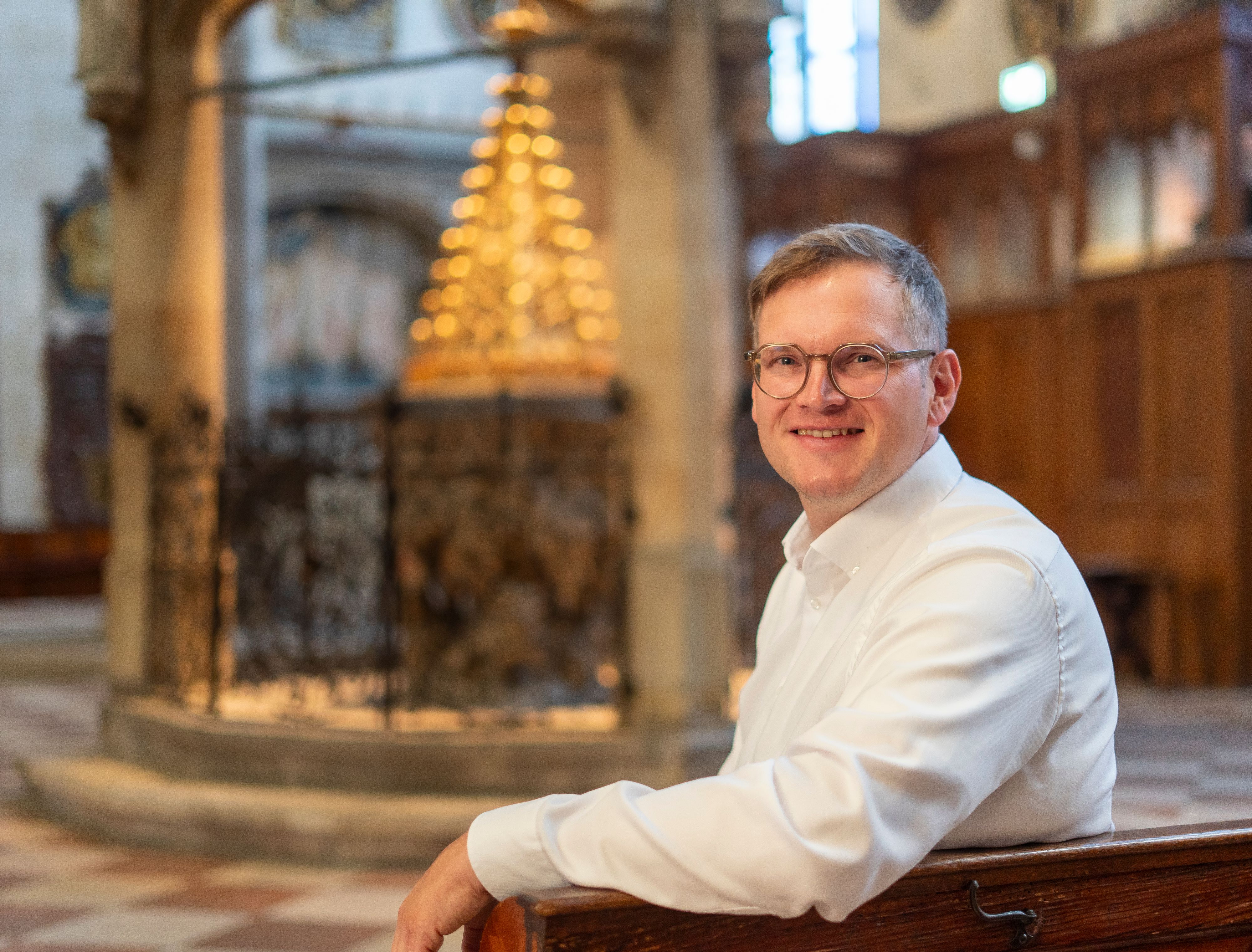 Julian Scharpf sitzt auf einer Kirchenbank. Im Hintergrund ist eine Kirche von Innen zu sehen. Der junge Mann trägt eine Brille, ein weißes Hemd und einen Kurzhaarschnitt.